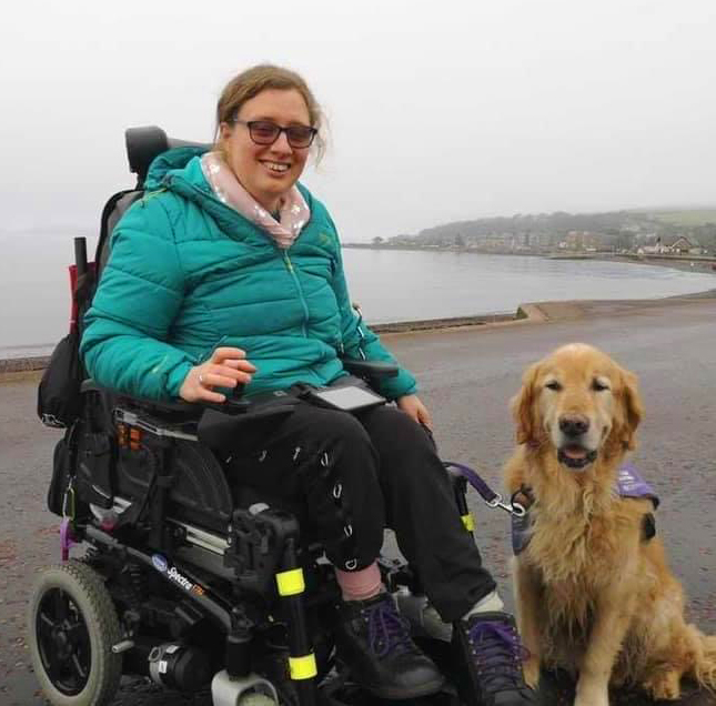 Payroll Service customer enjoying time at the beach. Picture is of a smiling lady and dog at the beach. The lady is using a powered wheelchair.